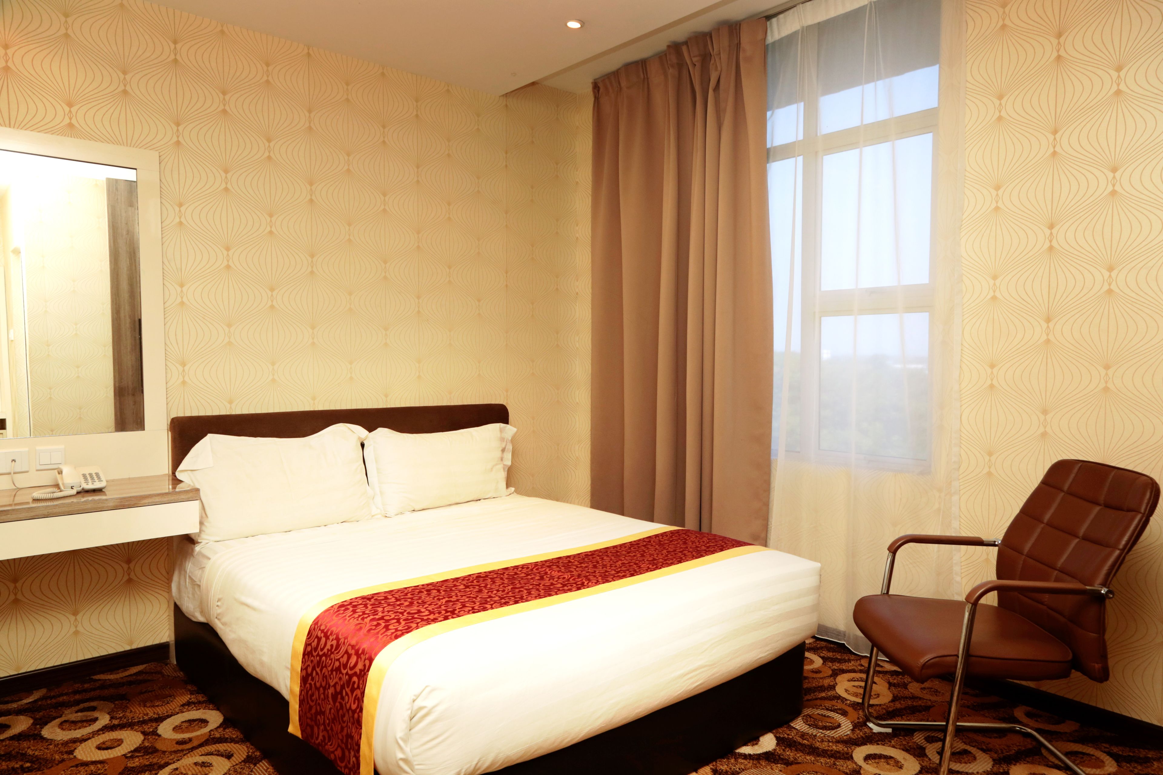 A hotel room with a bed, a chair, a desk with a telephone, and a window with curtains.