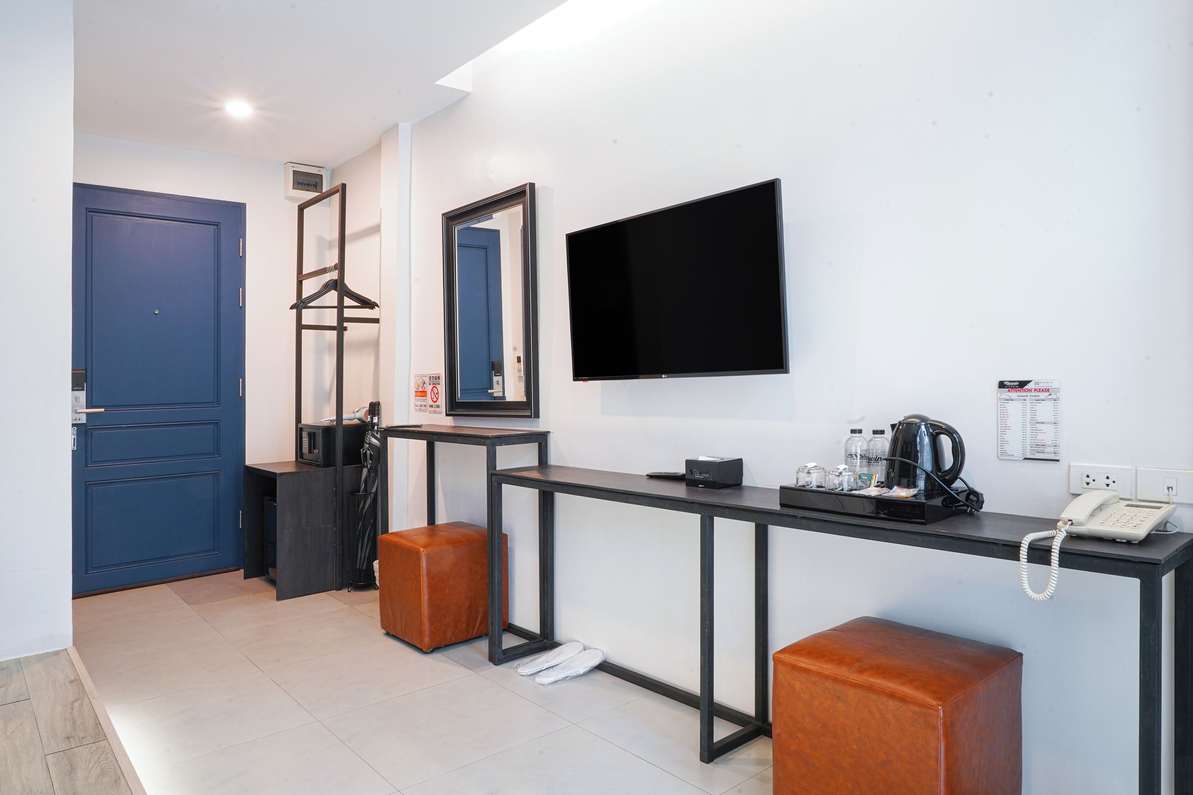A hotel room with a blue door, a black desk with a telephone, a mirror, and a television on the wall.