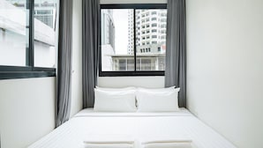 Ein Hotelzimmer mit Bett, weißen Laken, einem Fenster mit Vorhängen und Blick auf hohe Gebäude.
