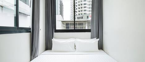 Quarto de hotel com cama, roupa de cama branca, janela com cortinas e vista para edifícios altos.