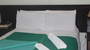Una cama bien hecha con almohadas blancas y una colcha verde, una toalla blanca doblada y un teléfono en la mesita de noche.