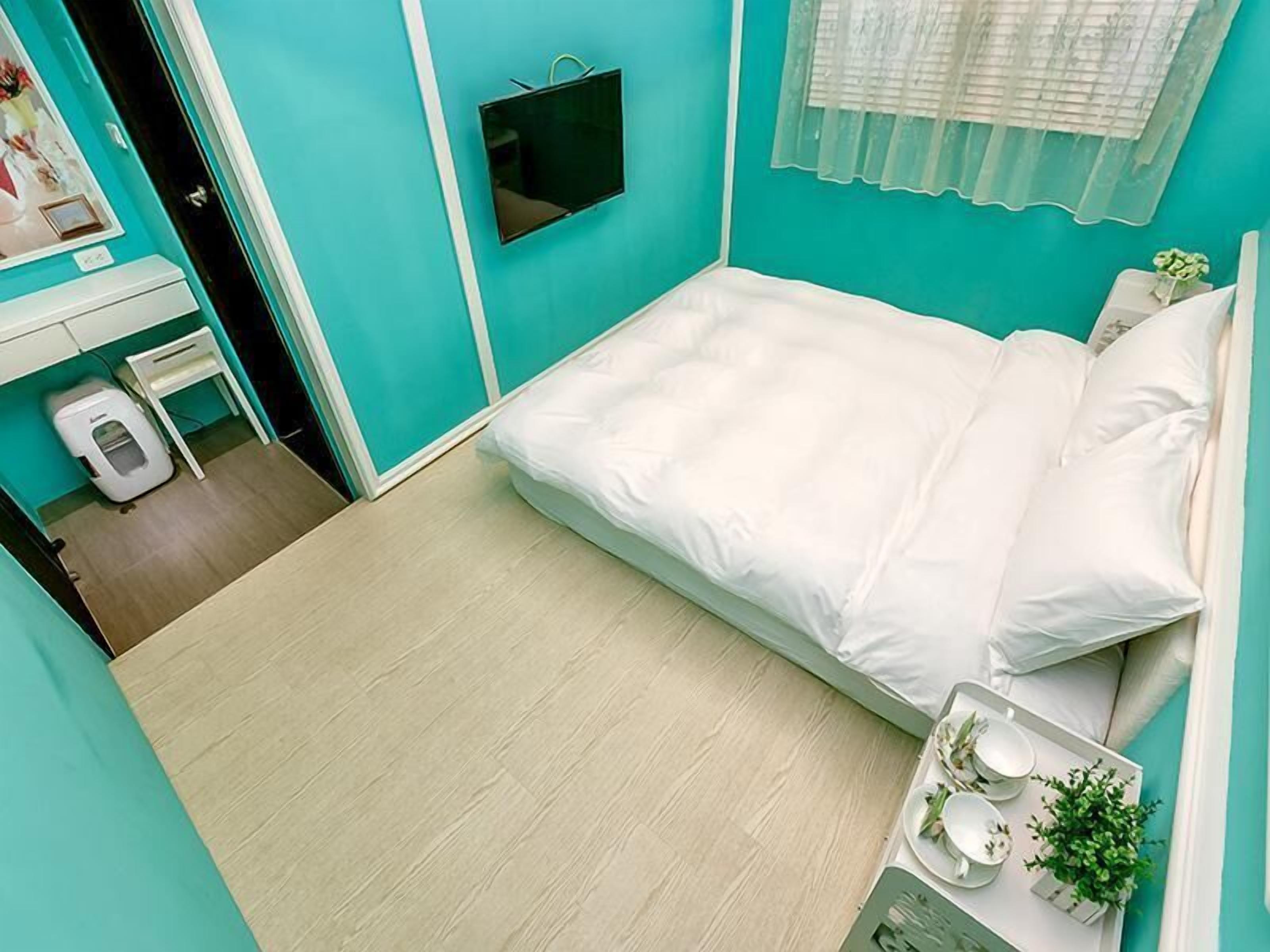 Un dormitorio con cama, una mesita de noche con una bandeja de objetos, un televisor montado en la pared y una ventana con cortina translúcida.