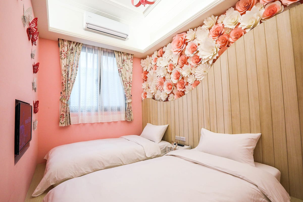 A bedroom with two beds, floral wall decor, a window with floral curtains, and an air conditioning unit.