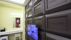 Een kamer met een aan de muur bevestigde televisie, een kleine witte stoel, een bureau en een plank met verschillende vakken, waarop 'Chocolade' staat.