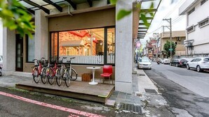 Een modern gebouw met een overdekt terras, fietsen die buiten geparkeerd staan, en een straatbeeld met auto's en gebouwen op de achtergrond.