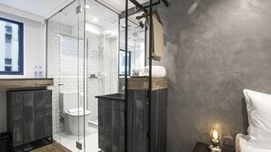 Un baño moderno con ducha envuelta en vidrio, tocador de madera y mesita de noche con lámpara.