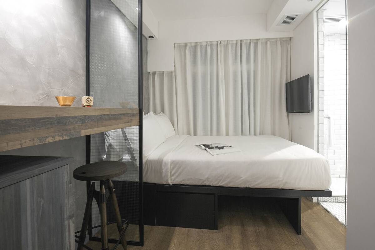 Una habitación de hotel moderna con una cama, un taburete pequeño, un televisor de pantalla plana y una ventana con cortinas.