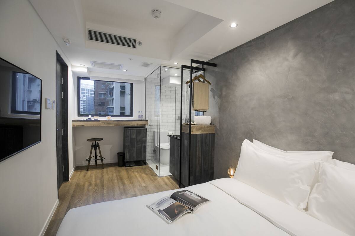 Habitación de hotel moderna con una cama grande, un escritorio pequeño, cabina de ducha de vidrio y vistas de la ciudad desde la ventana.