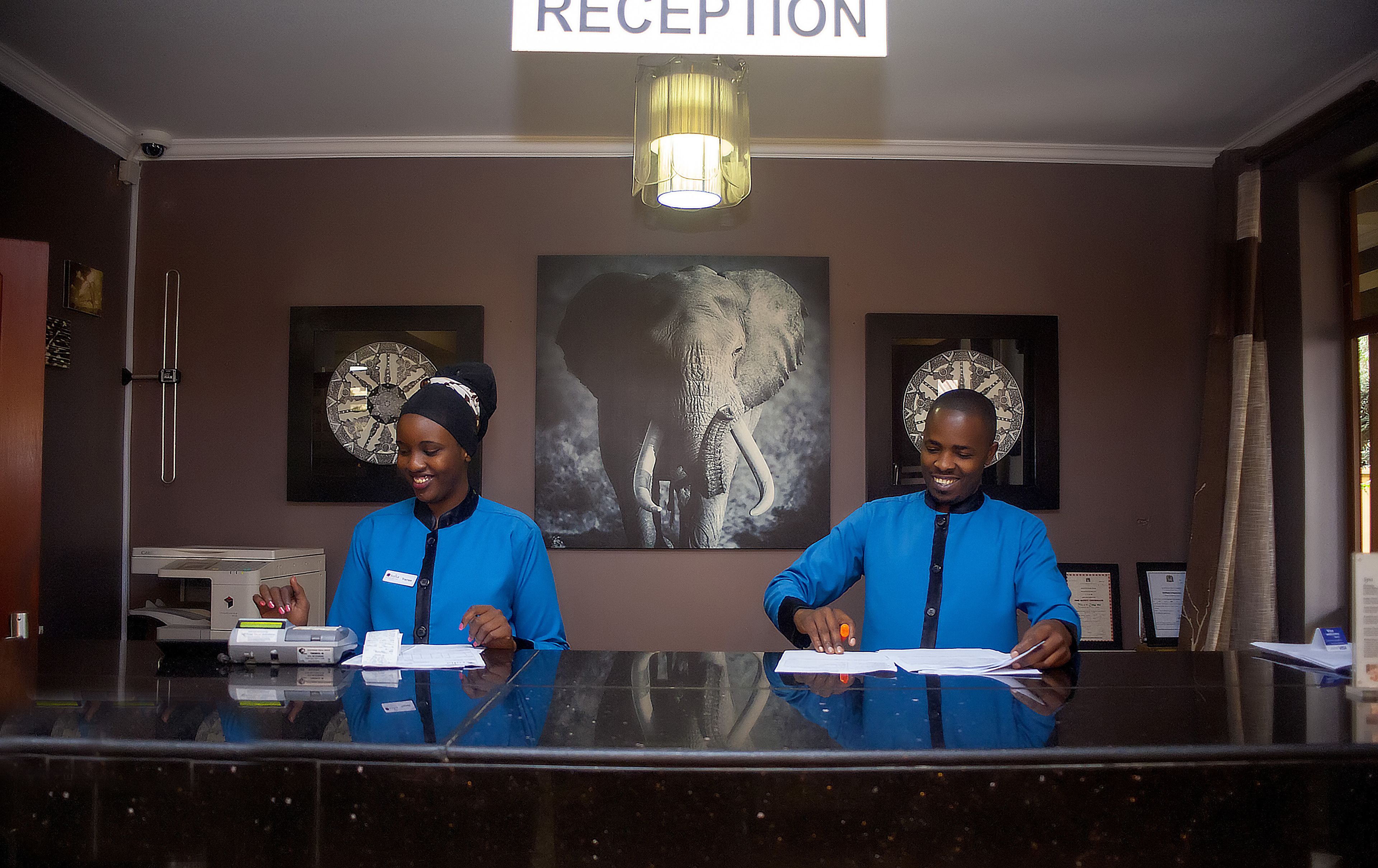 Un comptoir d’accueil avec deux employés, une enseigne indiquant « réception », et des œuvres d’art encadrées accrochées au mur.