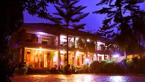 Un edificio de dos pisos con balcón, iluminado con luces cálidas, rodeado de plantas tropicales.