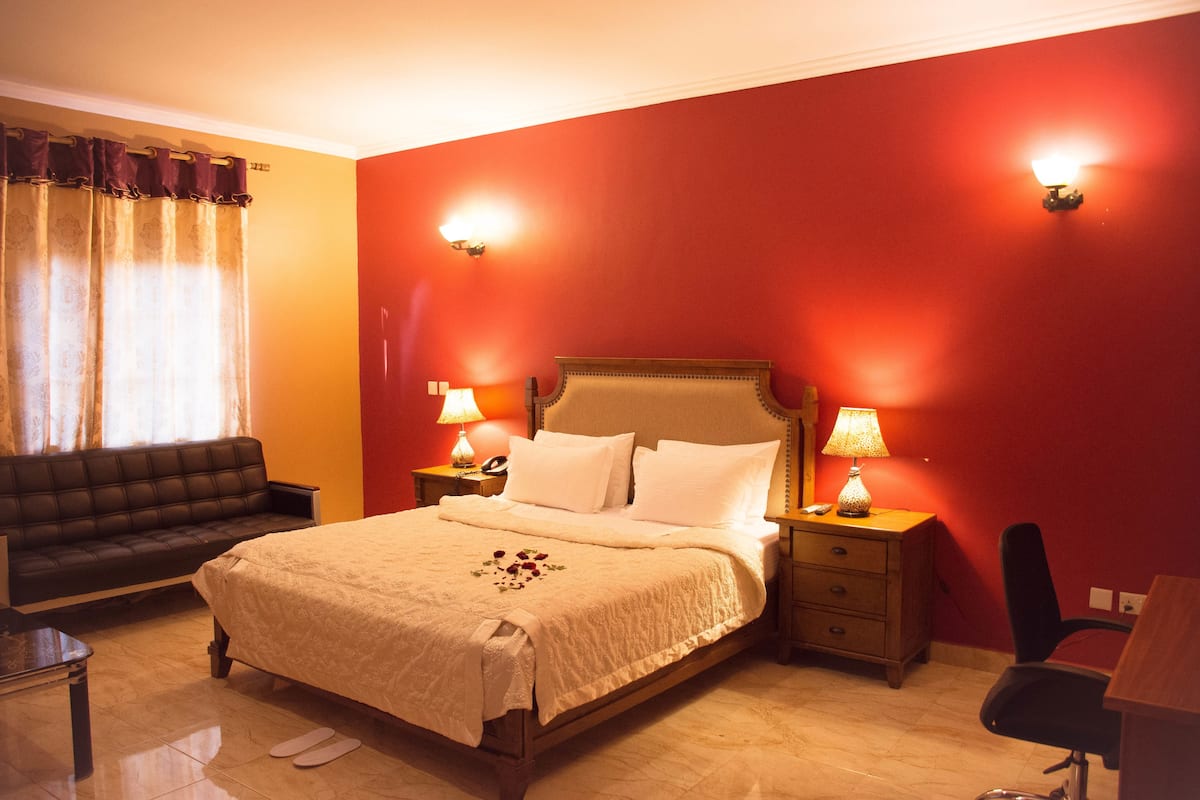 A hotel room with a large bed, two bedside tables with lamps, a wooden nightstand, a black leather sofa, and a wooden desk.