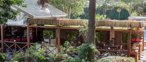 An outdoor restaurant with wooden tables and chairs, surrounded by lush greenery and a thatched roof structure.