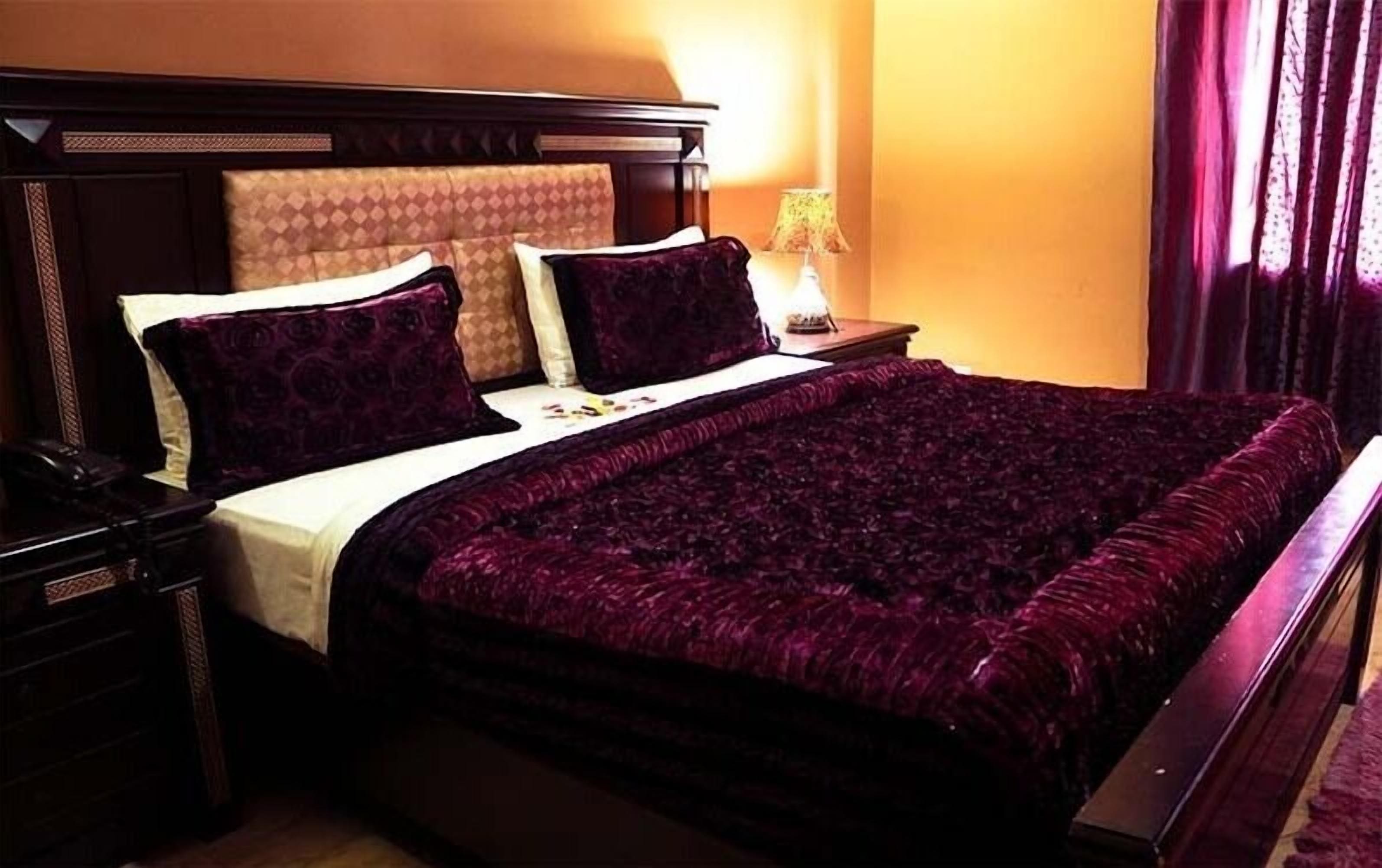 Un lit bien fait, avec une couvre-lit d’un violet foncé et des coussins assortis, une tête de lit en bois et une lampe de chevet.
