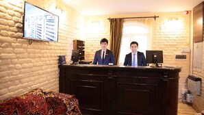 A reception desk with two staff members, a patterned sofa, a television mounted on a stone wall, and a window with curtains. - Star Hotel Tashkent (Tashkent)