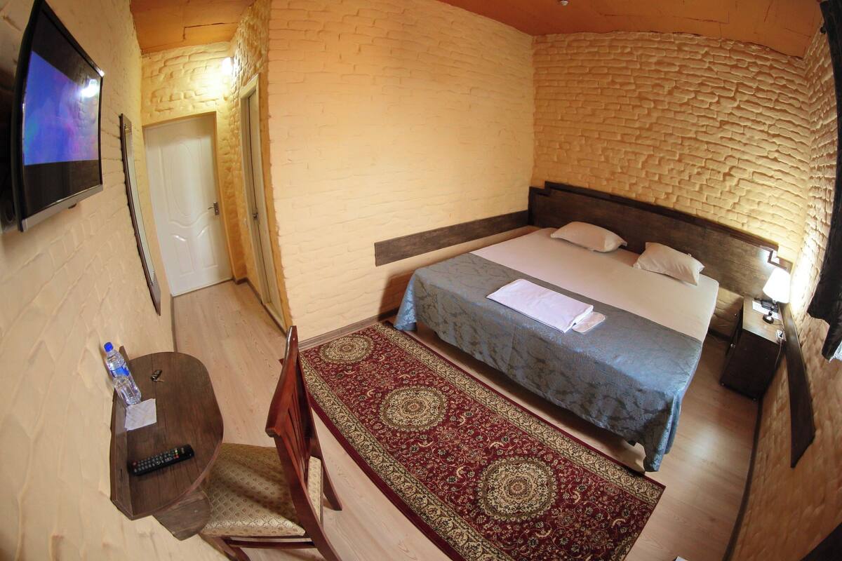 A bedroom with a bed, a TV mounted on the wall, a wooden table with a remote control, and a patterned rug.