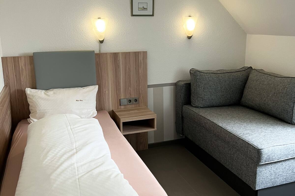 A modern hotel room with a bed, a nightstand, a sofa, and two wall-mounted lights.