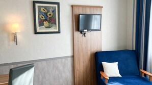 A hotel room with a blue sofa, a small table with a chair, a TV mounted on a wooden panel, and a framed floral artwork on the wall. - Hotel Garni Illertal (Altenstadt)