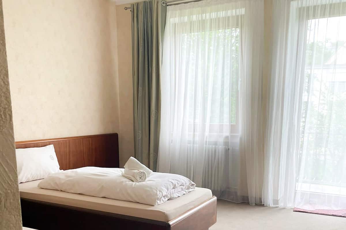 A hotel room with a double bed, a smaller bed, a wooden headboard, a large window with sheer curtains, and a carpeted floor.