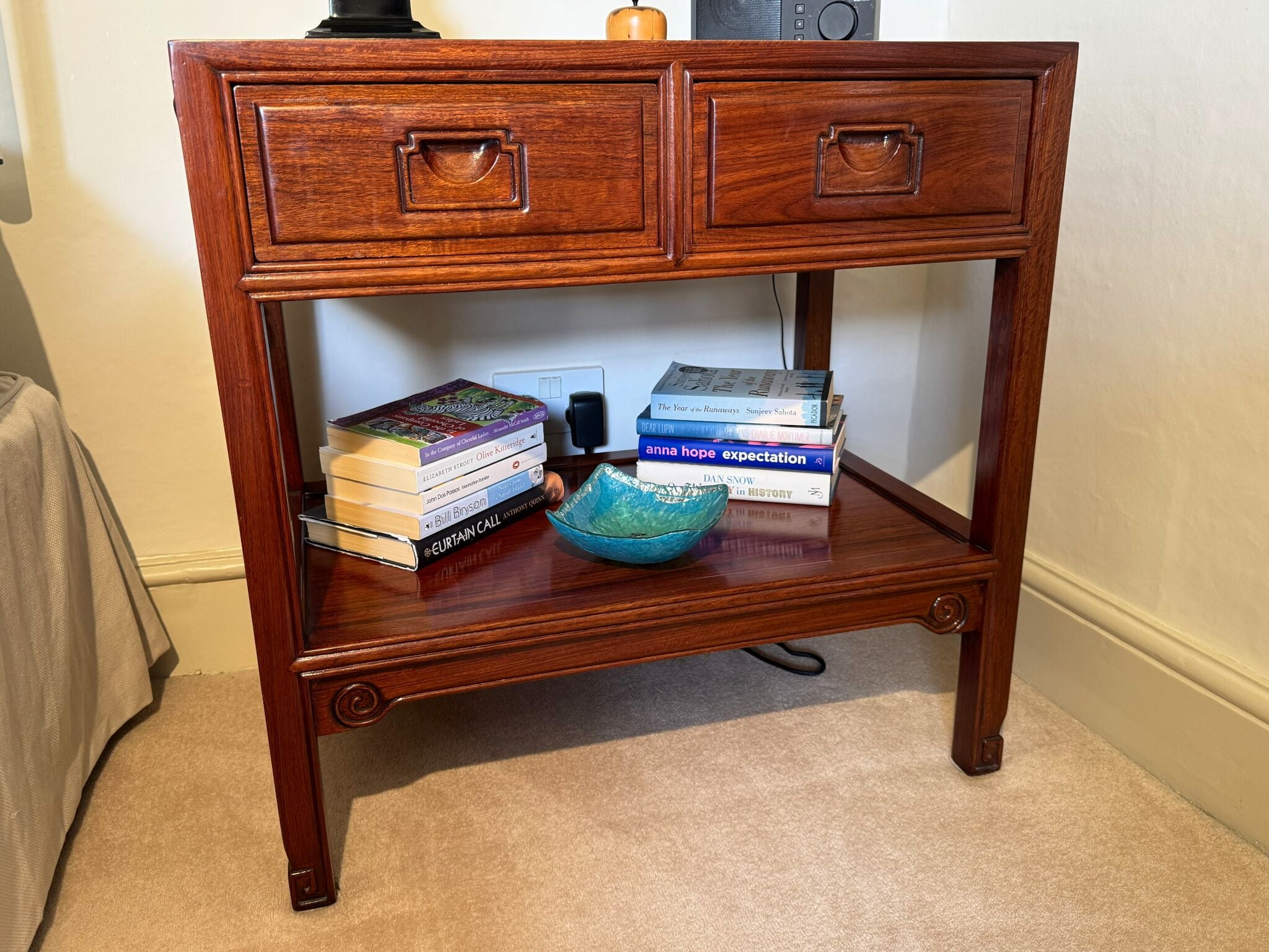 A wooden side table with two drawers and a shelf, holding books and a blue ceramic bowl.