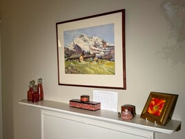 Un quadro incorniciato raffigurante un paesaggio montuoso su una parete, uno scaffale bianco con oggetti decorativi e un quadro con motivo floreale.
