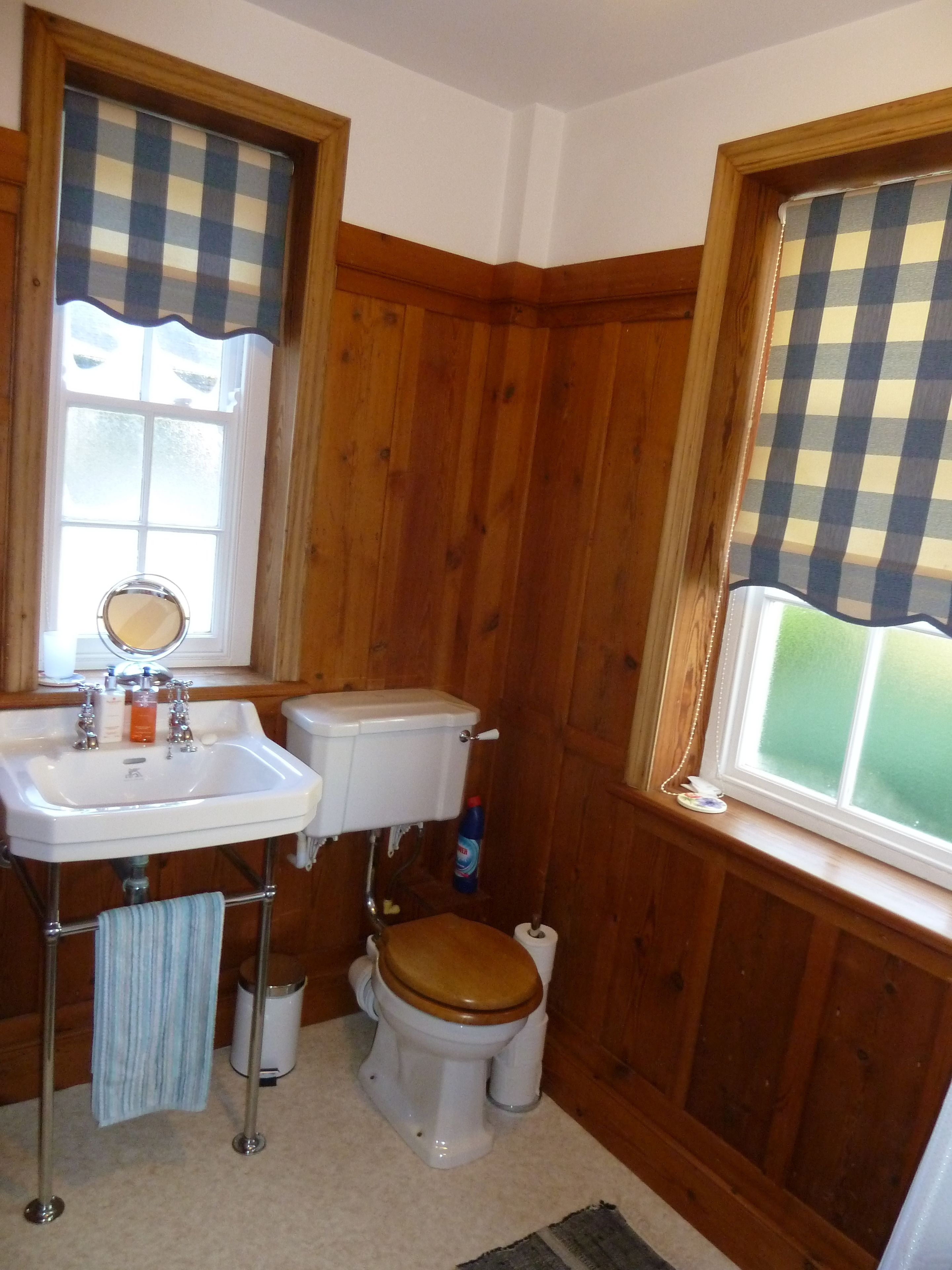 Un baño con paneles de madera, un lavabo blanco con grifería cromada, un inodoro con asiento de madera y una ventana con cortina de cuadros.