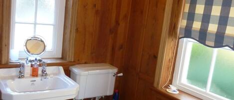 Un baño con paneles de madera, un lavabo blanco con grifería cromada, un inodoro con asiento de madera y una ventana con cortina de cuadros.