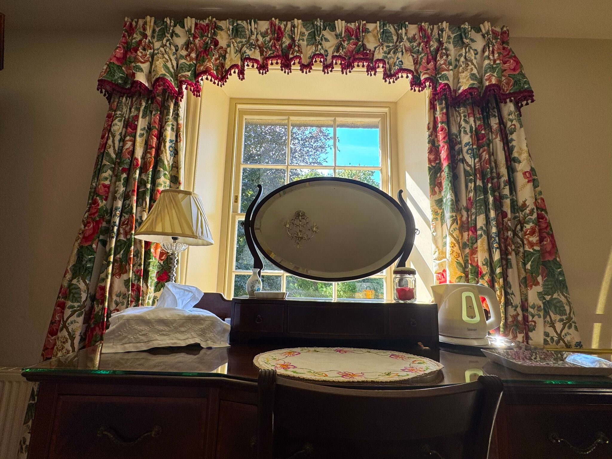 A room with a bed, a bedside table with a lamp, a mirror, and a window with curtains.