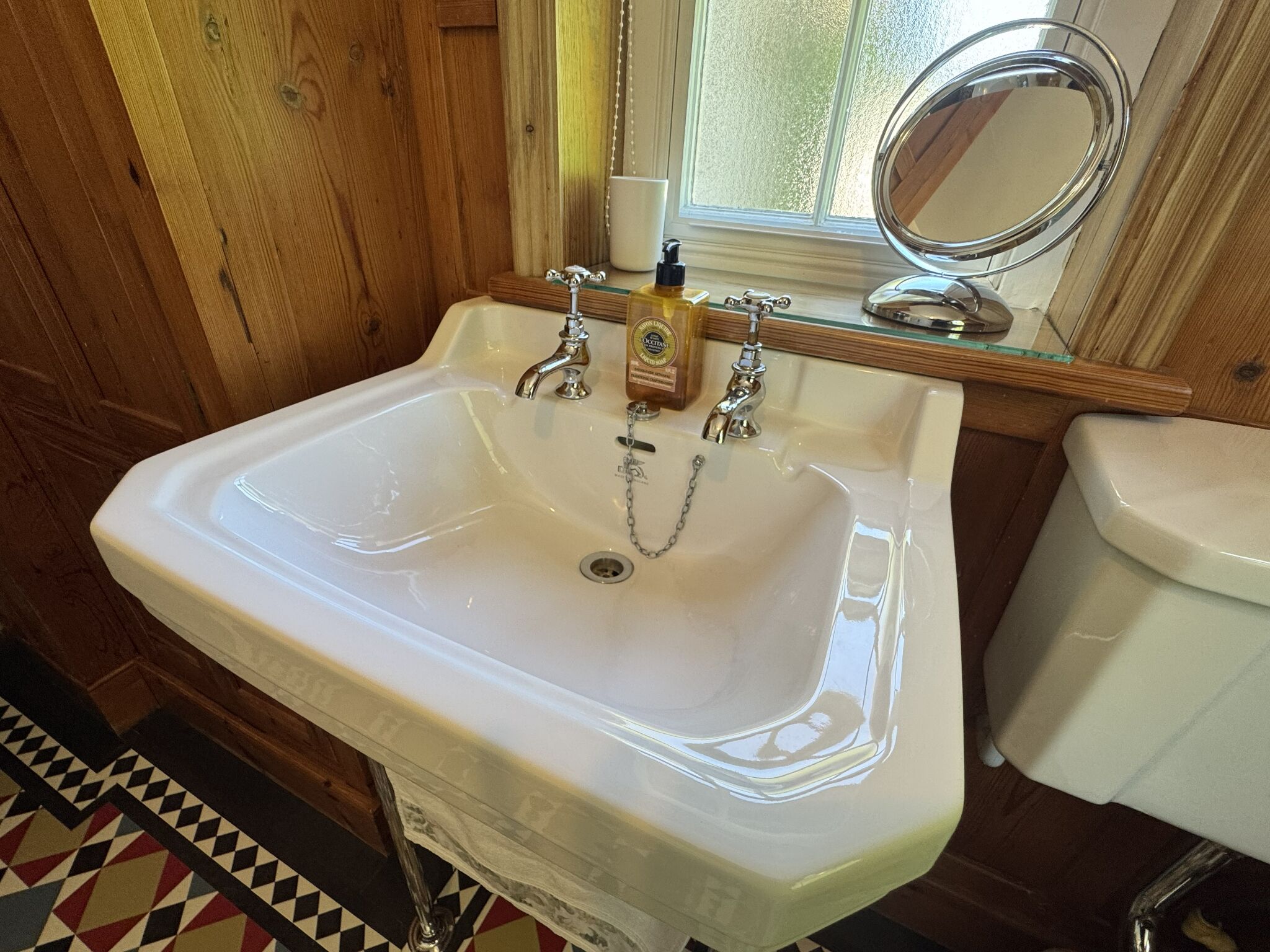 Un baño con un lavamanos blanco, dos grifos, un dispensador de jabón y un inodoro.