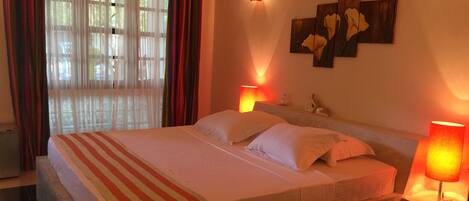 A hotel room with a large bed, two bedside lamps, a wall-mounted light, and a window with sheer curtains.