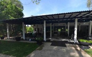 A covered patio area with a concrete floor, surrounded by greenery and a clear sky.