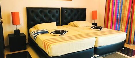 A double bed with a black headboard, two bedside tables with lamps, a colorful striped curtain, and a painting of elephants on the wall.