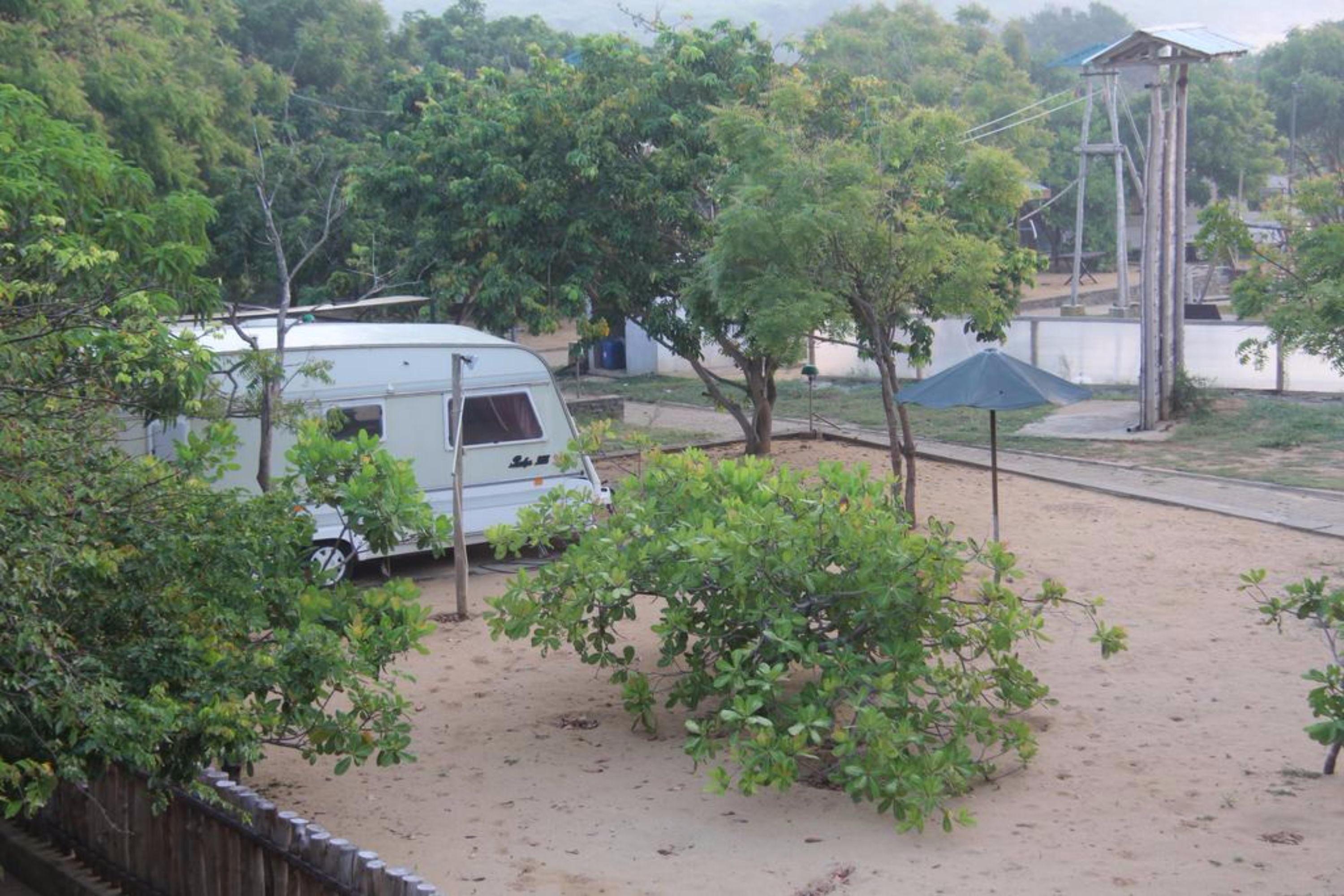 Una caravana blanca estacionada en una zona arenosa con árboles y una valla de madera.