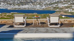 A poolside area with two lounge chairs and a small table, overlooking a coastal town with white buildings and a clear blue sea. - Nalu Villa Mykonos (Mykonos)