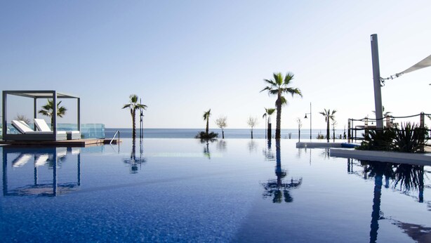 A clear blue infinity pool with a glass enclosure, palm trees, and a view of the ocean. - Sea Senses Apartments - Marholidays (Torrevieja)