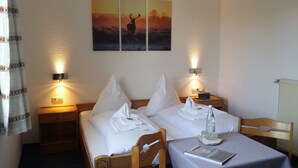 A hotel room with two beds, a table with a notebook, a bottle, and glasses. There are wall-mounted lights and a painting above the beds.