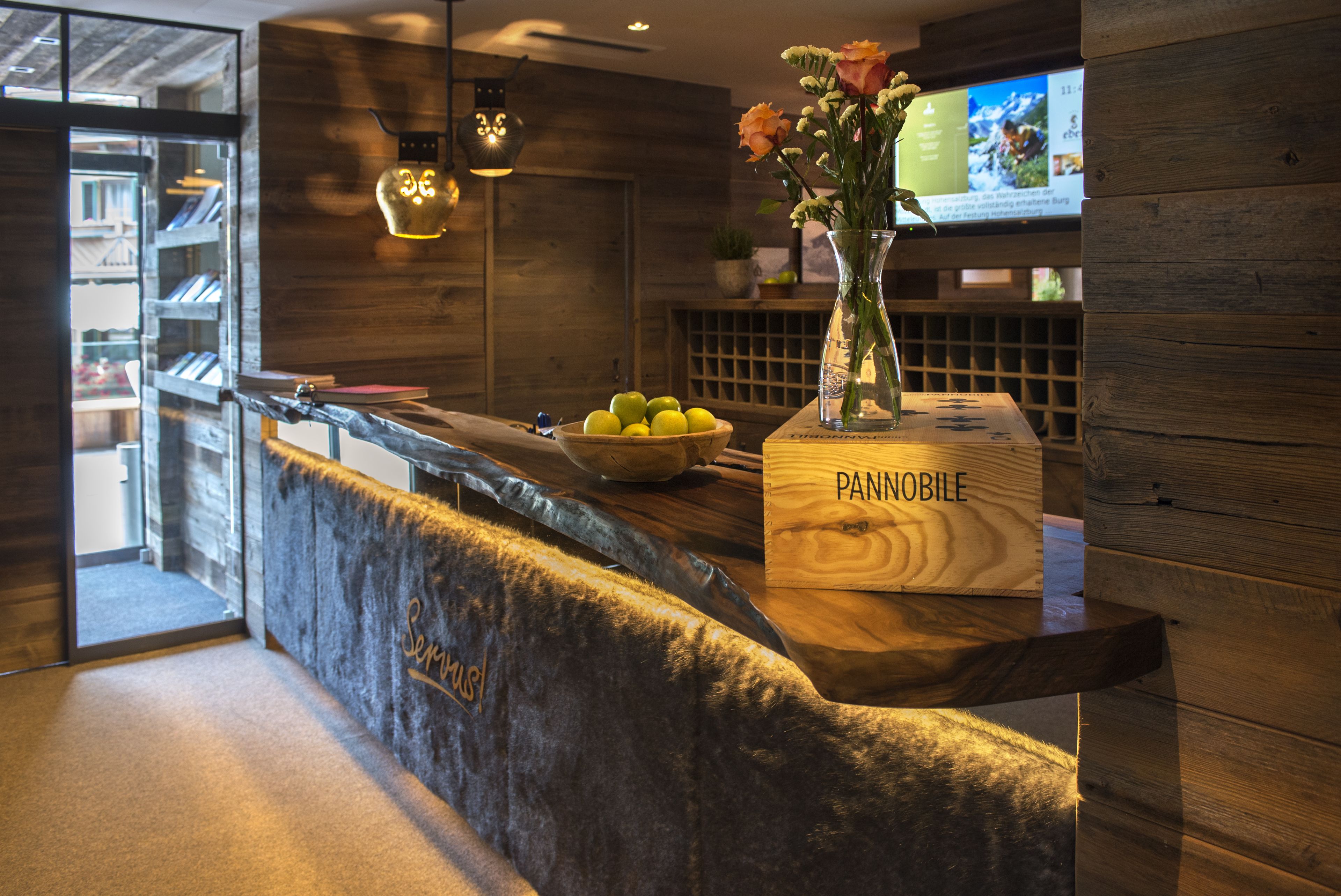 Una zona de recepció amb un taulell de fusta, un ram de flors, un plat de pomes i una pantalla digital amb informació.