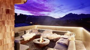 A cozy outdoor seating area with a fire pit, wooden benches, and a view of mountains under a twilight sky. - Lifestyle Hotel eder (Maria Alm am Steinernen Meer)
