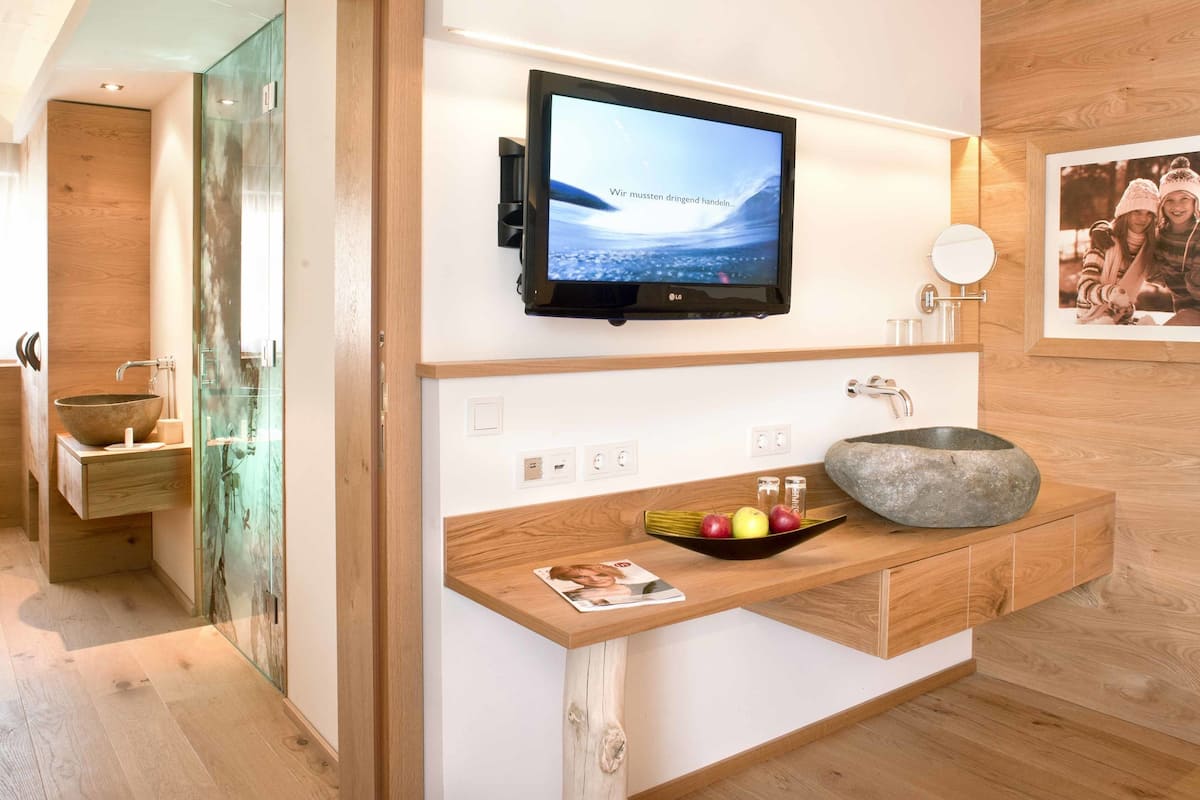 A modern bathroom with a wooden vanity, a stone sink, and a mounted TV displaying a message in German.