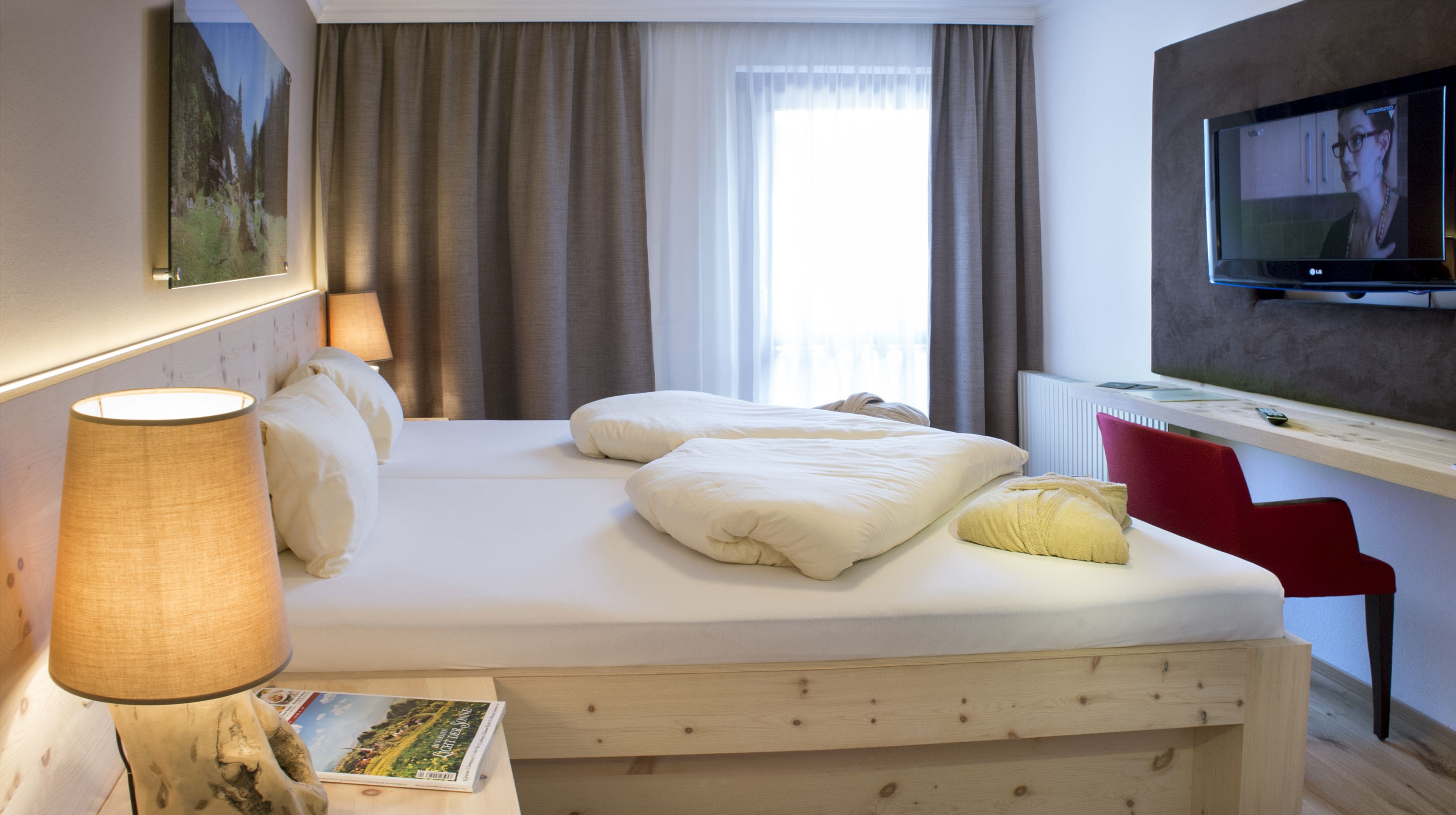 A hotel room with a bed, a desk with a TV, a lamp, and a window with curtains.