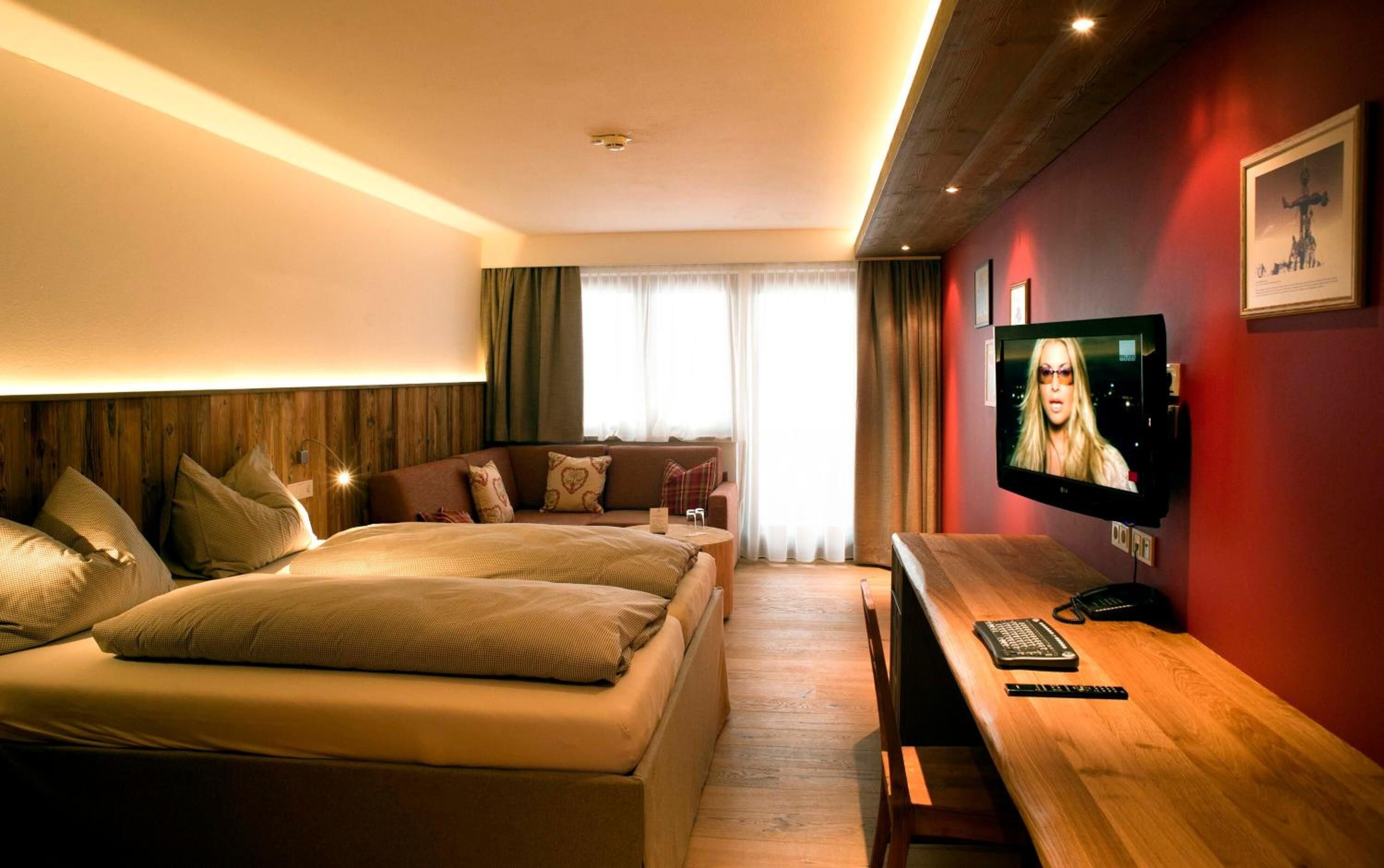 A hotel room with a wooden headboard, a sofa, a bed, a TV, and a wooden desk with a phone and remote control.