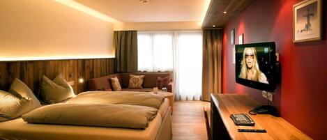 A hotel room with a wooden headboard, a sofa, a bed, a TV, and a wooden desk with a phone and remote control.