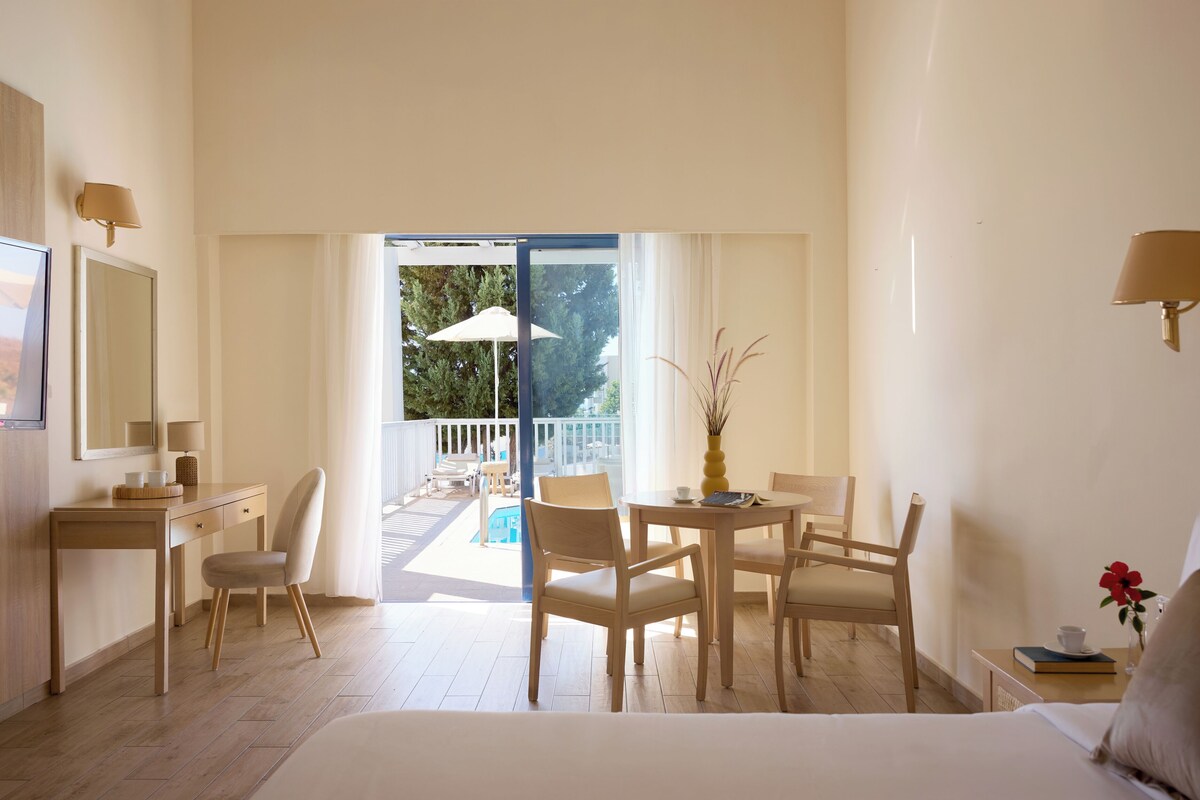 A hotel room with a bed, a desk, a chair, a table, and a balcony with a pool.