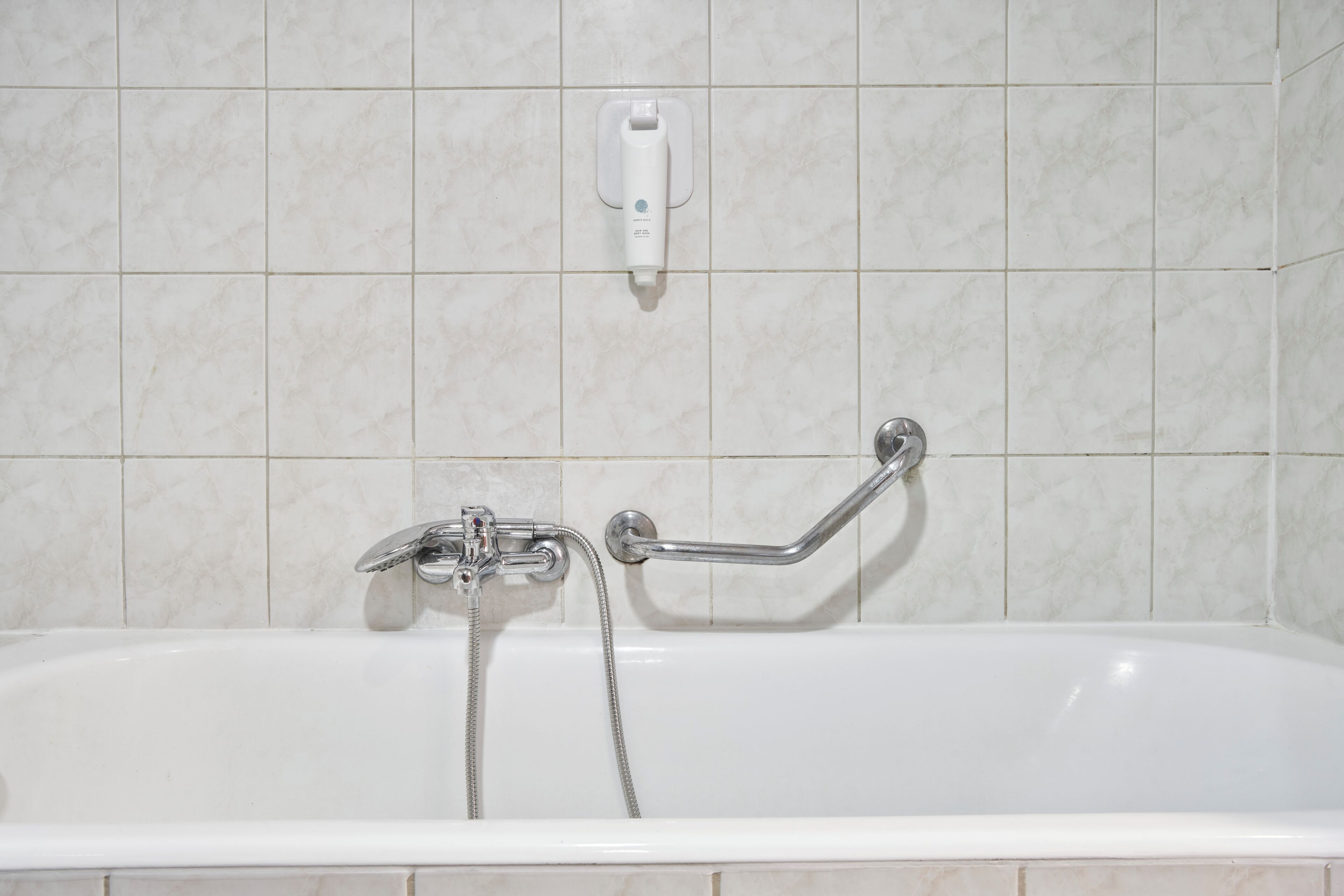 Una bañera blanca con ducha de mano y barra de apoyo, frente a una pared azulejada.