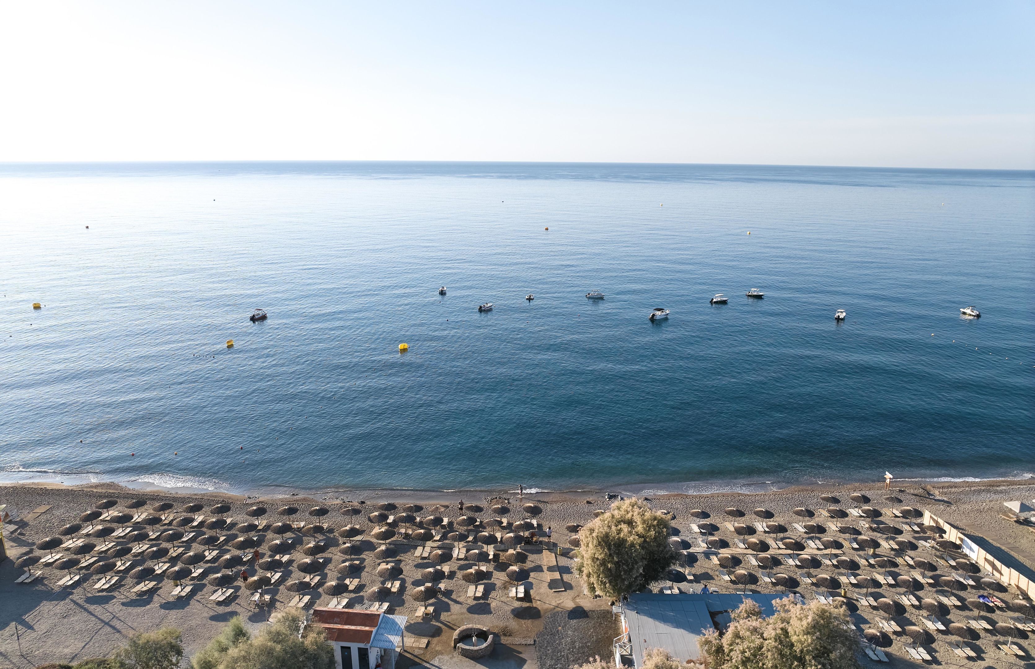 A beach with rows of umbrellas and sun loungers, a clear sky, and several boats in the water.