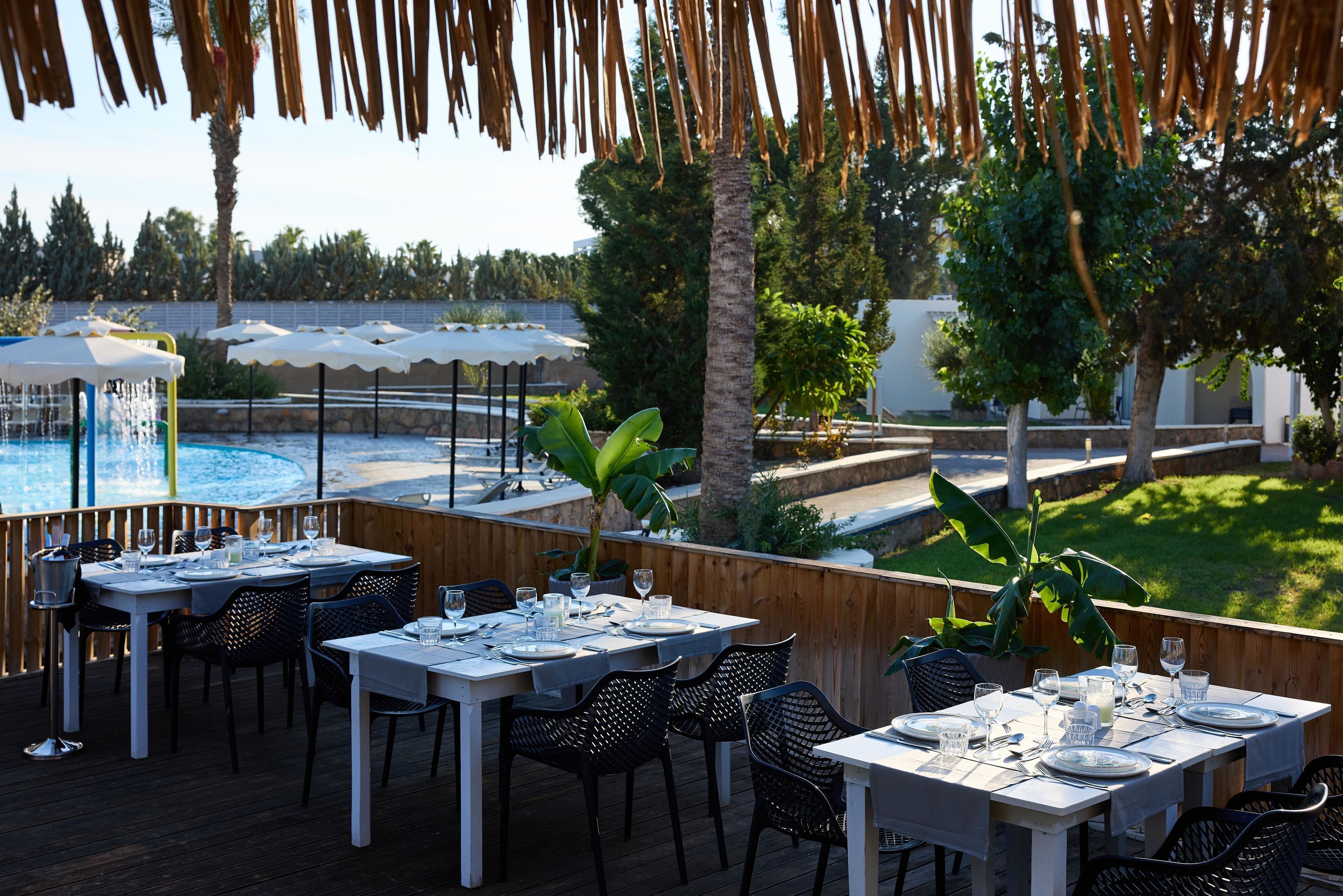 Un espace de restauration en plein air avec des tables pour deux, offrant une vue sur une piscine et un jardin aménagé.