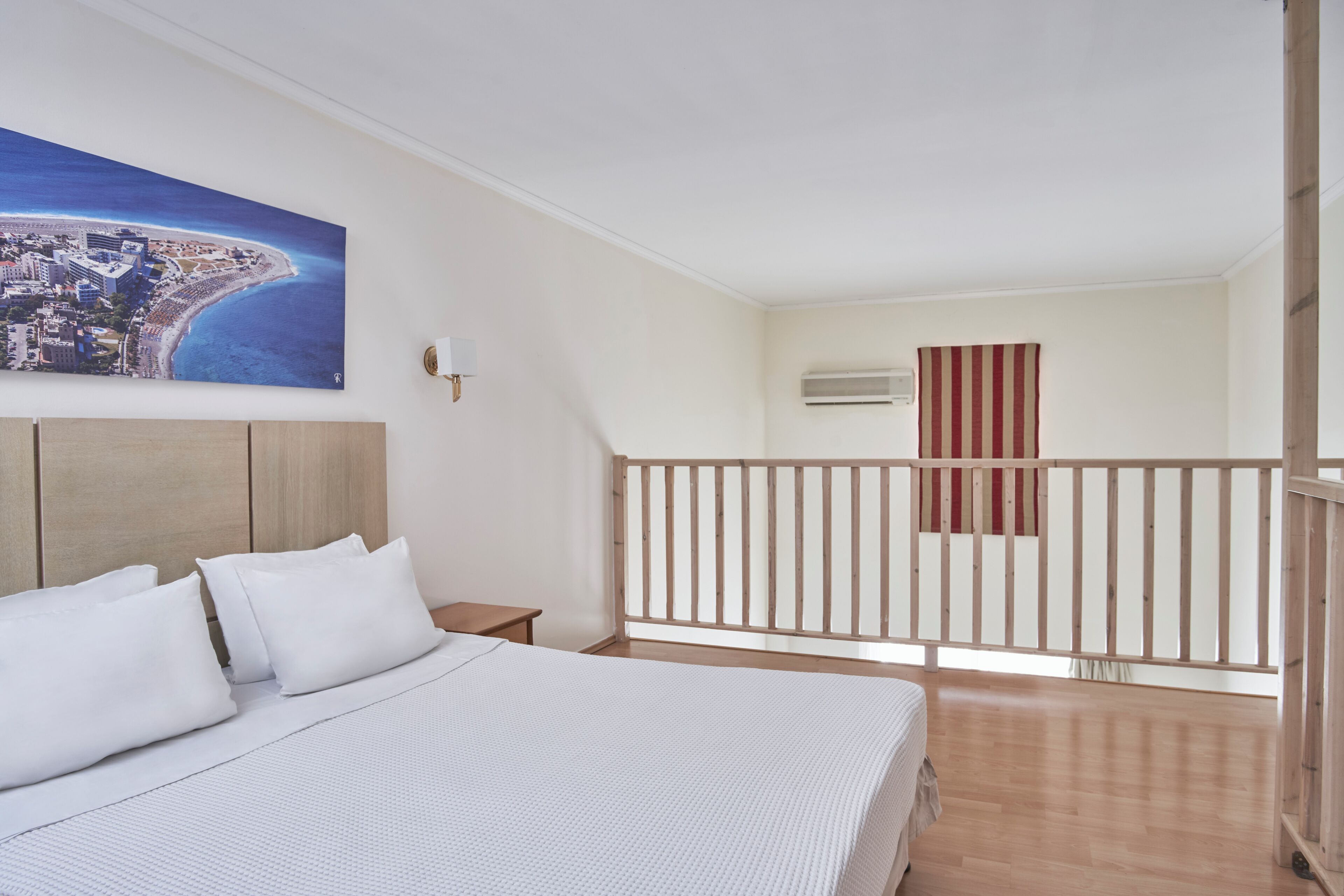 A bedroom with a bed, a nightstand, a painting of a coastal city, a wooden railing, and an air conditioning unit.