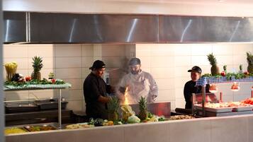 A kitchen with chefs preparing food, a buffet with various dishes, and a stainless steel hood above the cooking area.