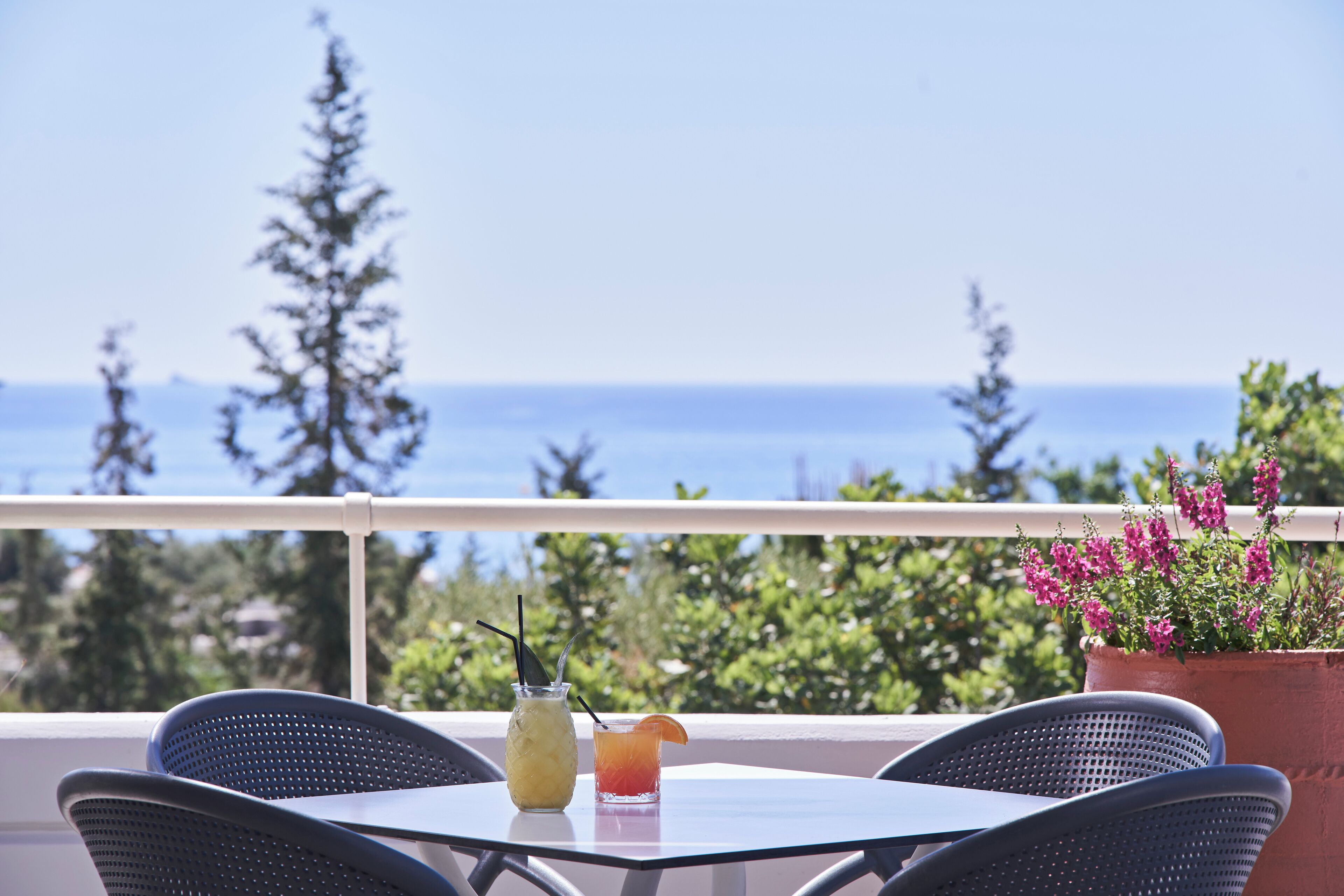 A balcony with a table, two chairs, a glass of a yellow drink, a glass of a red drink, and a potted plant with pink flowers.
