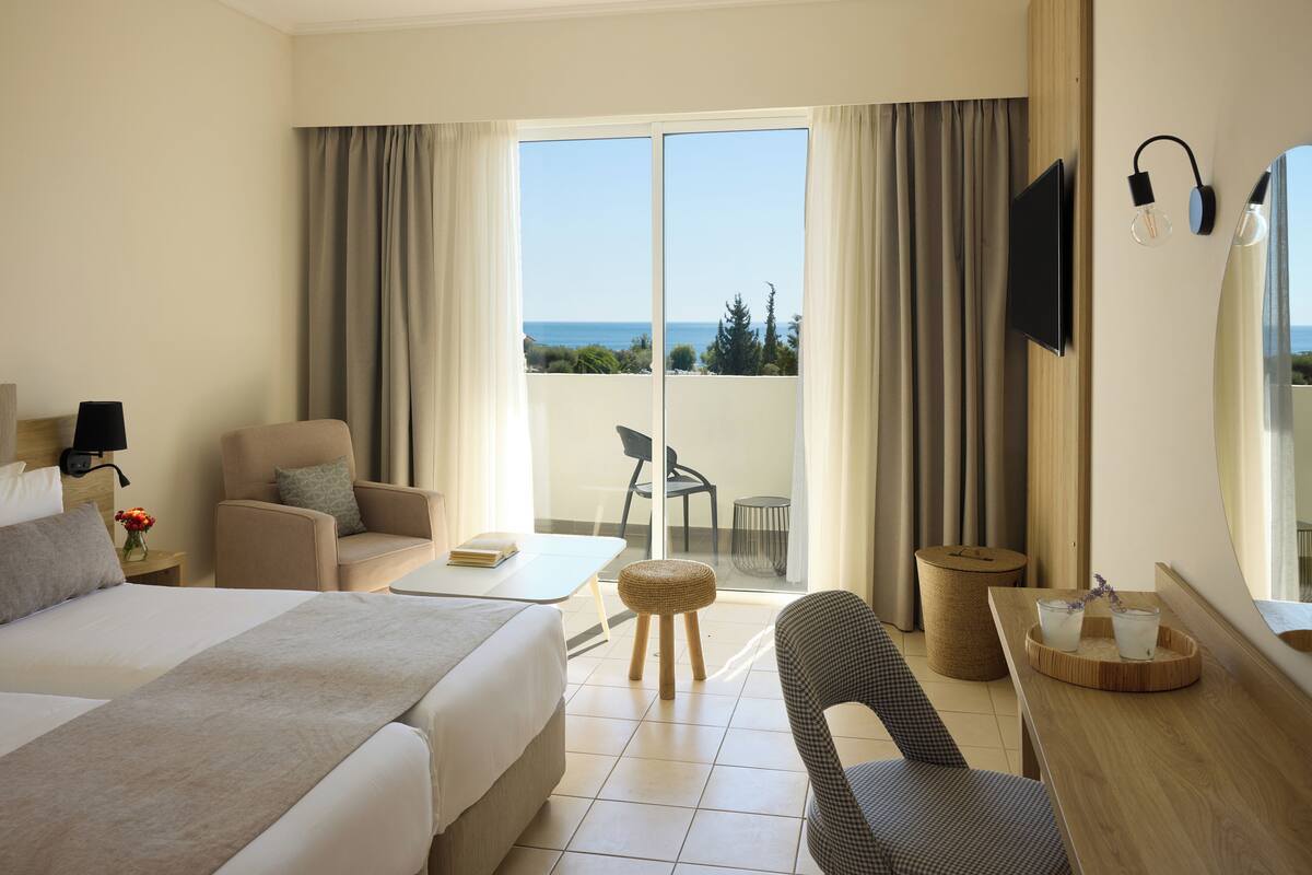Une chambre d’hôtel avec un lit, une chaise, une petite table, un téléviseur et une vue sur la mer.