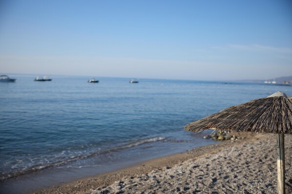 A beach with a straw umbrella, calm sea, and boats in the distance.